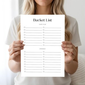 Person holding a bucket list planner page with two columns of checklist lines and small circle icons in a bright neutral indoor setting.
