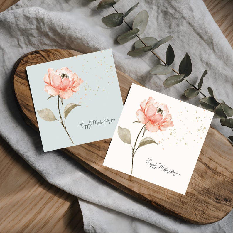 Two square floral greeting cards displayed on a wooden board, each featuring a soft watercolor pink flower with delicate gold speckles and script text.