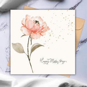 Square floral greeting card with a soft pink watercolor flower and gold accent details, displayed on a styled surface with a neutral envelope behind it.