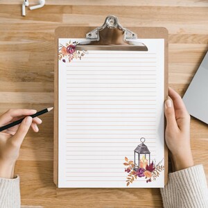 Fall stationery page with lined paper and lantern floral design on a clipboard, shown being written on by hand at a wooden desk. Cozy autumn theme digital download for home printing.