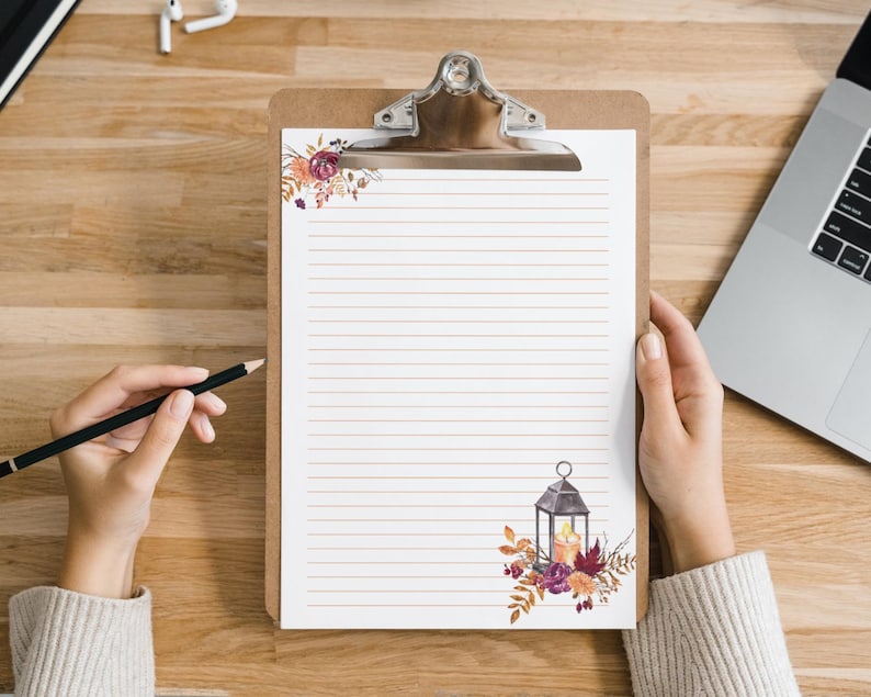 Fall stationery page with lined paper and lantern floral design on a clipboard, shown being written on by hand at a wooden desk. Cozy autumn theme digital download for home printing.