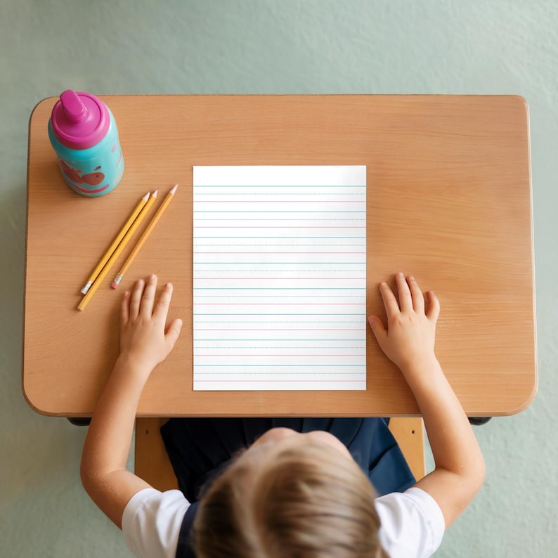Child sitting at a school desk with handwriting practice paper, pencils, and a water bottle in a classroom setting.