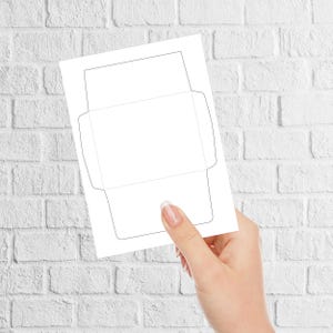 Envelope template held by a hand in front of a white brick background, showing solid cut lines and dotted fold lines on a plain sheet.