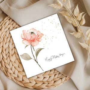 Square Mother’s Day card with watercolor pink flower and subtle gold speckles styled on woven rattan and neutral fabric background.