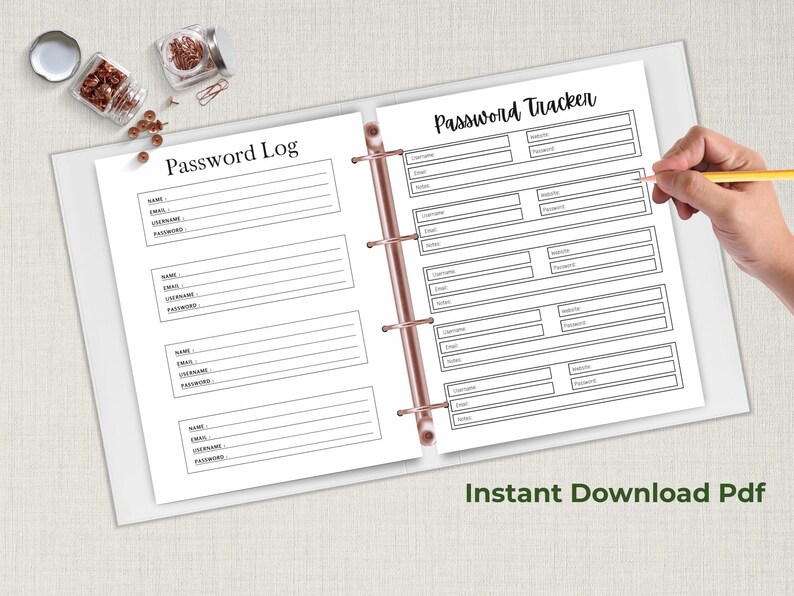 Open ring binder on a neutral fabric background showing two password organizer pages, one labeled Password Log and one labeled Password Tracker, with a hand writing on the page and small jars nearby.