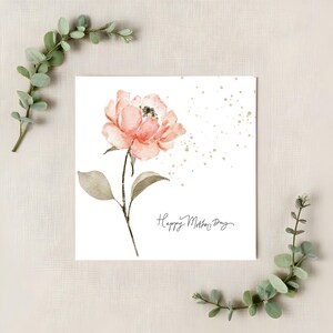 Square Mother’s Day card with watercolor blush flower and gold speckles styled on neutral linen background with greenery accents.
