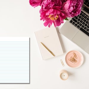 Lined notebook paper with blue horizontal lines and a red margin displayed on a white desk beside a laptop, notebook, gold pen, candle, and pink flowers.
