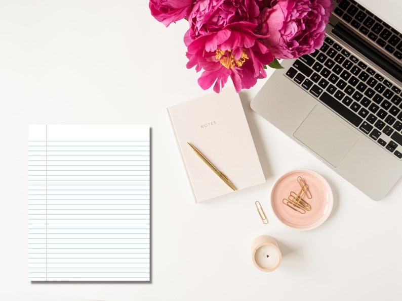 Lined notebook paper with blue horizontal lines and a red margin displayed on a white desk beside a laptop, notebook, gold pen, candle, and pink flowers.