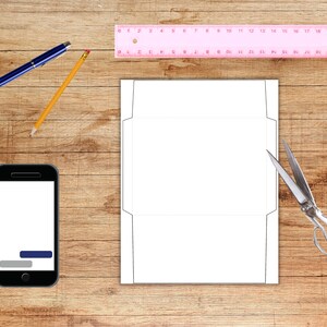 Envelope template sheet displayed on a woodgrain surface with crafting tools, including a pink ruler, pencil, pen, scissors, and a phone.