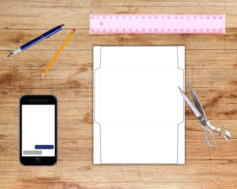 Envelope template sheet displayed on a woodgrain surface with crafting tools, including a pink ruler, pencil, pen, scissors, and a phone.