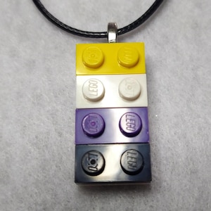 May include: A necklace featuring a pendant made of stacked LEGO bricks in yellow, white, purple, and black. The pendant is attached to a black cord with a silver-colored clasp. The LEGO bricks are arranged in a vertical column.