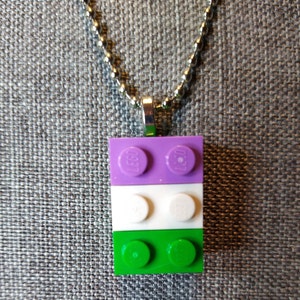 Op de afbeelding: Een zilveren ketting met een hanger gemaakt van drie gestapelde LEGO-stenen. De bovenste steen is paars, de middelste steen is wit en de onderste steen is groen. De LEGO-stenen hebben het LEGO-logo erop gedrukt.