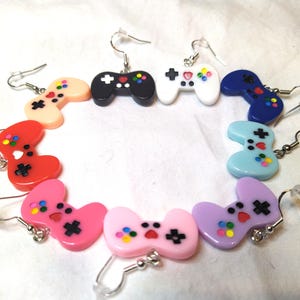 May include: A collection of colorful, novelty earrings shaped like video game controllers. The earrings are in various colors, including black, white, pink, red, blue, and purple, with colorful button details and silver-tone ear wires.