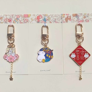 May include: Three gold keychains with different charms. The first charm is a pink and white gift box with a small rabbit and cherry blossoms. The second charm is a blue and white circle with a white rabbit and fireworks. The third charm is a red diamond with two white rabbits and Chinese characters.