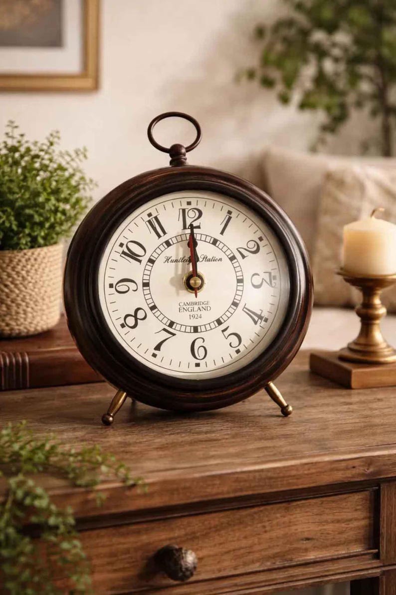 May include: A round, dark brown wooden table clock with a cream-colored face, black Arabic numerals, and a metal ring handle on top. The clock face features the text Huntley Station, Cambridge England, and 1924, with thin black hands and a red second hand.