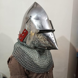May include: A shiny silver medieval helmet with a pointed crown and a visor. The helmet features red leather trim with rivets and a chainmail neck guard. The visor has small holes. A historical replica.