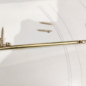 Könnte beinhalten: Ein goldfarbener Metallzirkel mit einem langen, geraden Arm und einem kürzeren, abgewinkelten Arm. Der Zirkel hat an einem Ende eine scharfe Spitze und am anderen Ende eine Bleistiftspitze. Der Zirkel ist zum Zeichnen von Kreisen und Bögen bestimmt.