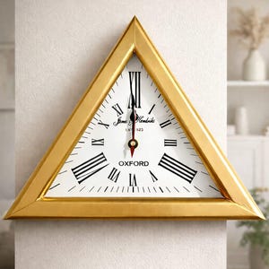 Puede incluir: Un reloj de pared triangular con marco dorado y esfera blanca. El reloj presenta números romanos negros, manecillas negras y el texto "OXFORD". También está presente el texto "James & Hendricks EST 1923".