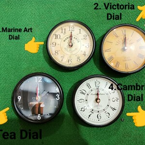 May include: Four round wall clocks with black frames. Clock 1, Marine Art Dial, has a cream face with an anchor. Clock 2, Victoria Dial, features a yellow face with Roman numerals. Clock 3, Tea Dial, shows a cup graphic. Clock 4, Cambridge Dial, has a white face.