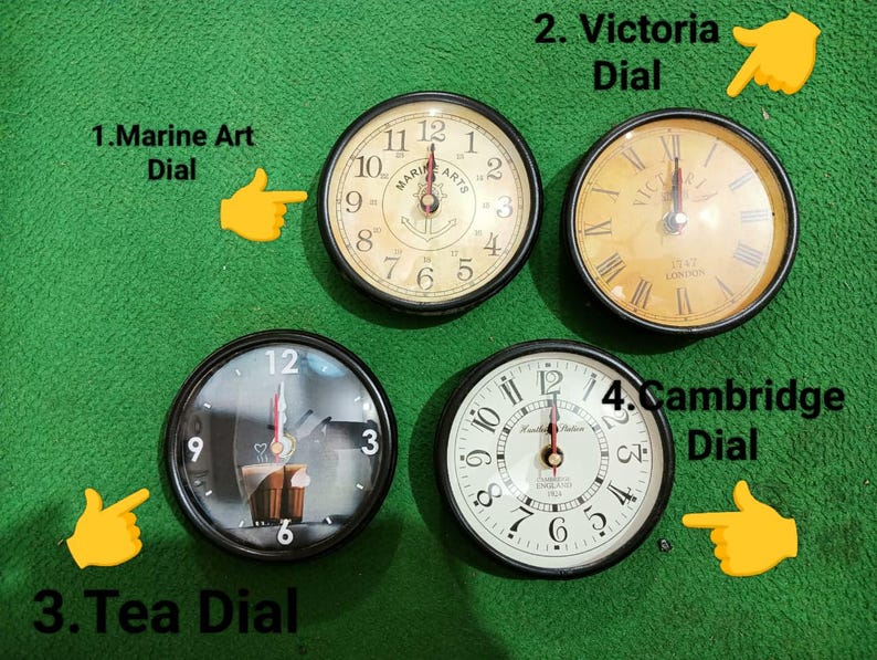 May include: Four round wall clocks with black frames. Clock 1, Marine Art Dial, has a cream face with an anchor. Clock 2, Victoria Dial, features a yellow face with Roman numerals. Clock 3, Tea Dial, shows a cup graphic. Clock 4, Cambridge Dial, has a white face.