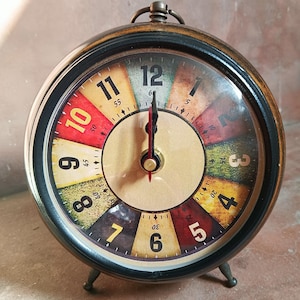 May include: A round analog alarm clock with a dark metal frame and a multi-colored face. The dial features large black numerals from 1 to 12, with each hour segment colored in shades of red, yellow, green, and brown. It has two black hands.