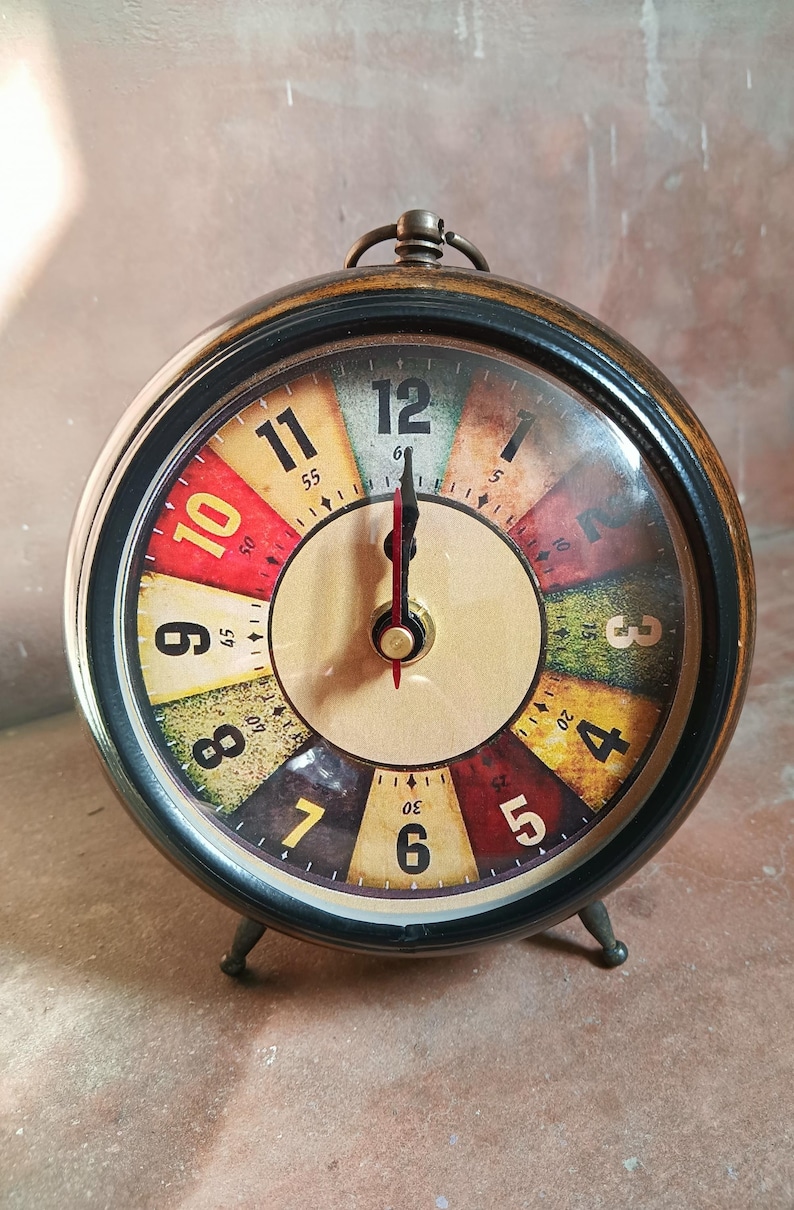May include: A round analog alarm clock with a dark metal frame and a multi-colored face. The dial features large black numerals from 1 to 12, with each hour segment colored in shades of red, yellow, green, and brown. It has two black hands.
