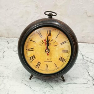 May include: A round table clock with a dark brown frame and a yellow, aged-look face. The clock features black Roman numerals, black hour and minute hands, a red second hand, and the text "VICTORIA" and "1747 LONDON" printed on the dial.