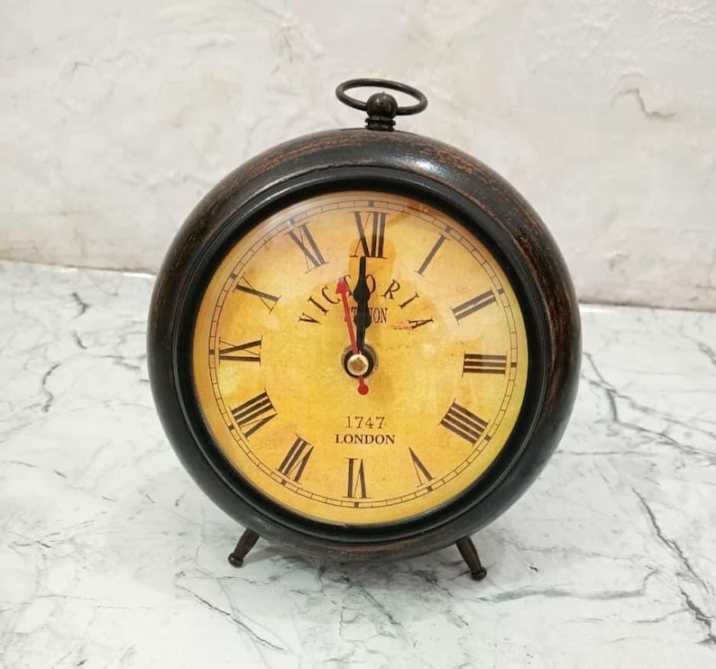 May include: A round table clock with a dark brown frame and a yellow, aged-look face. The clock features black Roman numerals, black hour and minute hands, a red second hand, and the text "VICTORIA" and "1747 LONDON" printed on the dial.