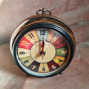 May include: A round analog table clock with a dark wood-toned frame and a metal ring handle on top. The clock face features a multi-colored segmented background with black numerals from 1 to 12 and smaller minute markers, plus red hands.