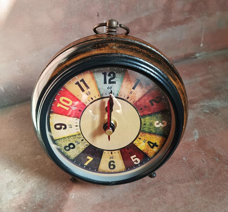 May include: A round analog table clock with a dark wood-toned frame and a metal ring handle on top. The clock face features a multi-colored segmented background with black numerals from 1 to 12 and smaller minute markers, plus red hands.