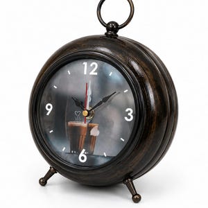 May include: A round, dark brown, distressed-finish metal alarm clock with a top ring handle and two small feet. The clock face features white numbers 3, 6, 9, and 12, black hour and minute hands, a red second hand, and a graphic of coffee cups.