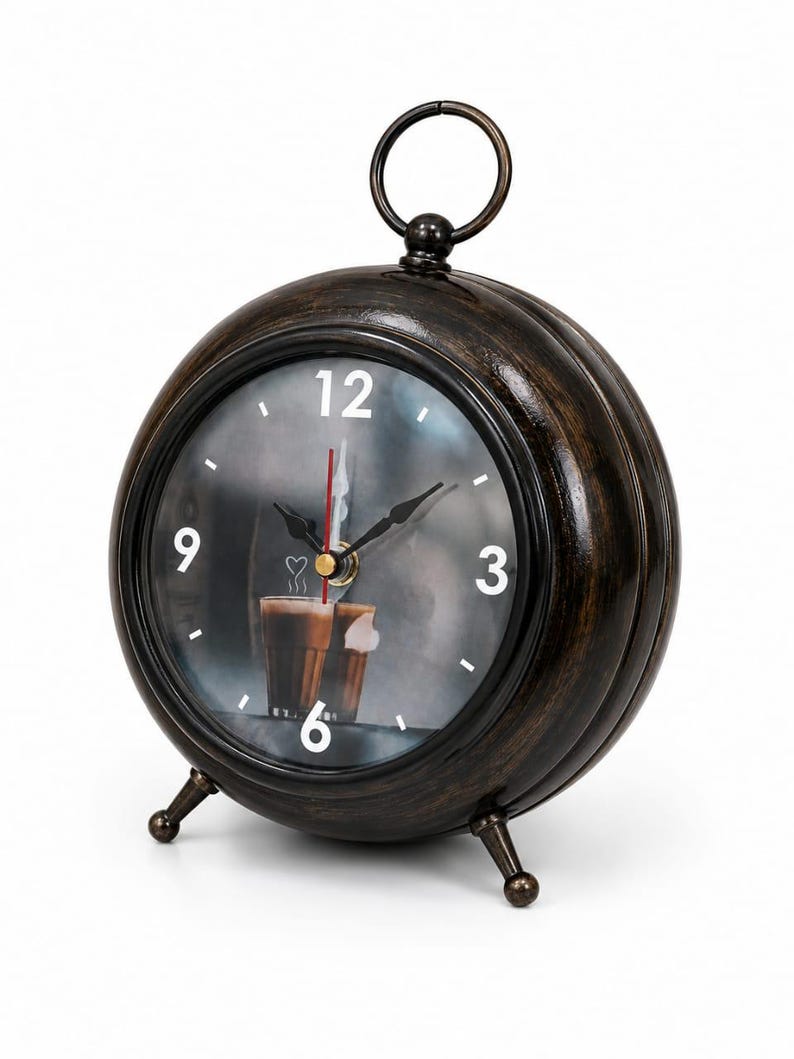 May include: A round, dark brown, distressed-finish metal alarm clock with a top ring handle and two small feet. The clock face features white numbers 3, 6, 9, and 12, black hour and minute hands, a red second hand, and a graphic of coffee cups.