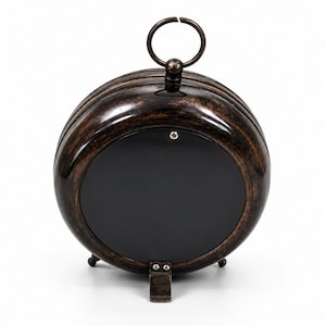 May include: A round, dark brown, metallic clock with a distressed finish. The back features a smooth, black circular panel secured by a single screw at the top. A metal ring handle sits at the apex, and a small, hinged metal stand is at the base.