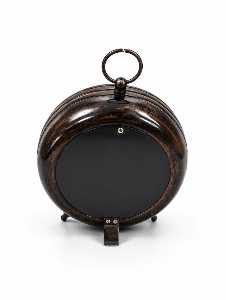 May include: A round, dark brown, metallic clock with a distressed finish. The back features a smooth, black circular panel secured by a single screw at the top. A metal ring handle sits at the apex, and a small, hinged metal stand is at the base.
