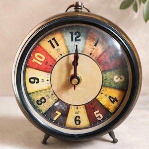 Könnte beinhalten: Vintage-Wecker mit farbenfrohem Zifferblatt mit den Zahlen 1 bis 12. Die Uhr hat eine schwarze Lünette und einen bronzefarbenen Rahmen. Die Zeiger zeigen auf 12 Uhr. Die Uhr wird von drei kleinen Beinen getragen.