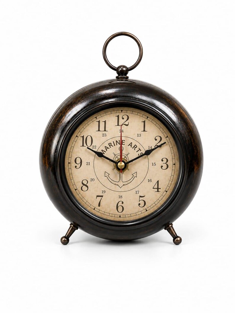 May include: A round, dark bronze-toned table clock with a ring handle on top and two small feet. The clock face is cream-colored with black numerals 1 through 12, smaller 13 through 24 markers, a central anchor graphic, and the text MARINE ARTS.