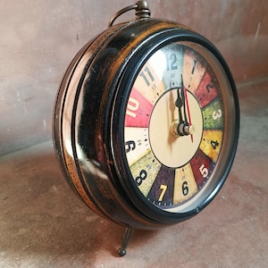May include: A round table clock with a dark, distressed wooden frame and a metal handle on top. The clock face features a colorful, segmented design with black numerals from 1 to 12, black hour and minute hands, and a thin red second hand.