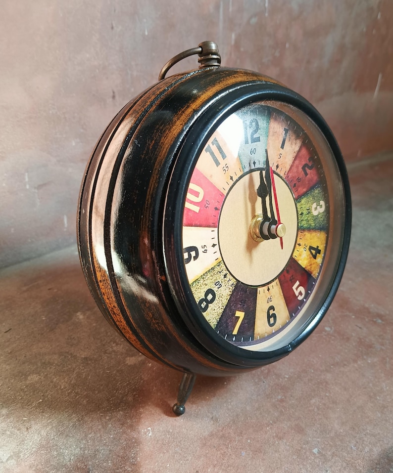 May include: A round table clock with a dark, distressed wooden frame and a metal handle on top. The clock face features a colorful, segmented design with black numerals from 1 to 12, black hour and minute hands, and a thin red second hand.