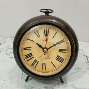 May include: A round, dark brown table clock with a metallic finish, featuring a cream-colored face with black Roman numerals. The clock displays the text Digo London and Royalty 1890. It has black hour and minute hands and a red second hand.