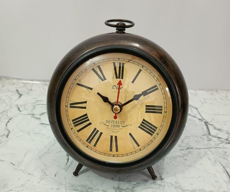 May include: A round, dark brown table clock with a metallic finish, featuring a cream-colored face with black Roman numerals. The clock displays the text Digo London and Royalty 1890. It has black hour and minute hands and a red second hand.