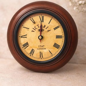 May include: A round, wooden clock with a dark brown finish. The clock face is cream-colored with black Roman numerals and hands. The text "VICTORIA STATION LONDON 1747" is printed on the face. The clock is approximately 5 inches in diameter.