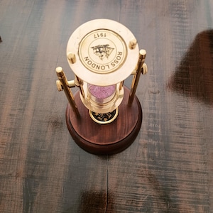 May include: A vintage hourglass with a brass frame and a dark wooden base. The top of the hourglass features the text "ROSS LONDON" and the year "1917". The sand inside is a pinkish color.