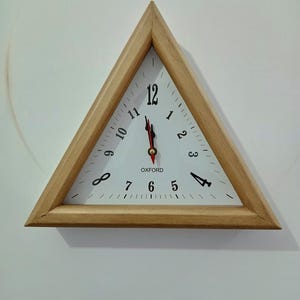 May include: A triangular wall clock with a light wood frame and a white face. The clock features black numerals from 1 to 12, with the word OXFORD printed in black below the center. It has black hour and minute hands and a red second hand.