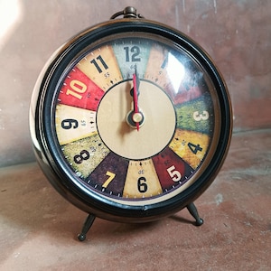 May include: A round analogue alarm clock with a black metal frame and two small legs. The clock face features a multi-coloured segmented background with black numerals from 3 to 12. It has black hour and minute hands and a thin red second hand.