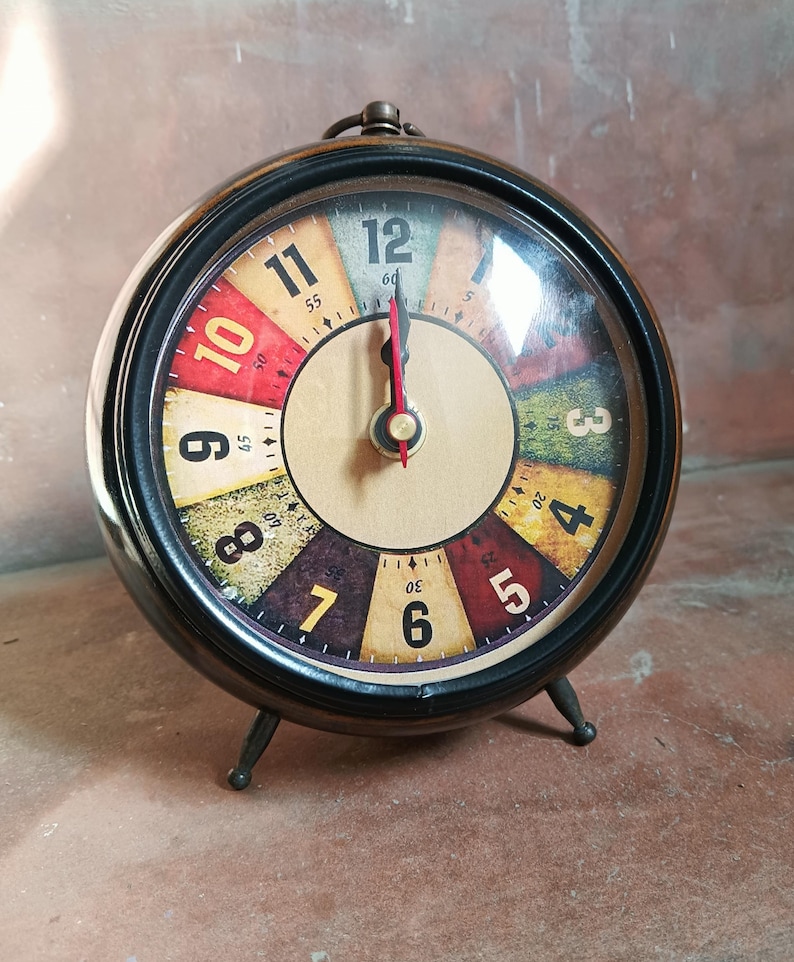 May include: A round analog alarm clock with a black metal frame and two small legs. The clock face features a multi-colored segmented background with black numerals from 3 to 12. It has black hour and minute hands and a thin red second hand.