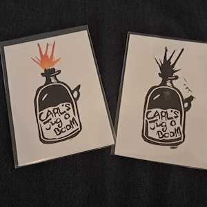 May include: Two rectangular prints on white paper, each encased in a clear plastic sleeve. The prints feature a black ink illustration of a jug with a fuse and an explosion at the top. The text "Carl's Jug o Boom" is printed on the jug.