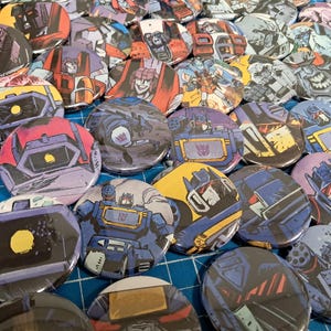 May include: A collection of colorful, round buttons featuring various Transformers characters in action poses. The buttons showcase a variety of colors, including red, blue, yellow, and purple, with detailed illustrations and designs.