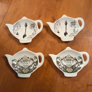 Könnte beinhalten: Vier weiße Keramik-Teekannen-Teetassenuntersetzer. Zwei haben ein schwarzes Löffeldesign mit Krone und dekorativen Schnörkeln. Die anderen beiden haben ein grün-graues Blumendesign mit dem Schriftzug "GREEN TEA".