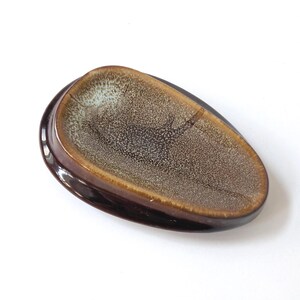May include: A brown ceramic dish with an unusual, organic shape. The dish has a dark brown exterior and a speckled, tan interior with a lighter rim. The design is reminiscent of a natural form, suitable for serving or display.