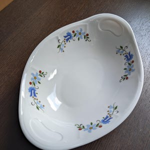 Puede incluir: Plato ovalado blanco para servir con un diseño floral. El plato presenta un borde con asa y está decorado con flores azules, hojas verdes y bayas rojas. El plato está sobre una superficie marrón oscuro.